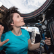 An astronaut holds up her hand in Star Trek's live long and prosper hand signal as she gazes out of her craft at the earth. She's wearing a plain blue t-shirt and her short hair is lifting upwards due to the lack of gravity.